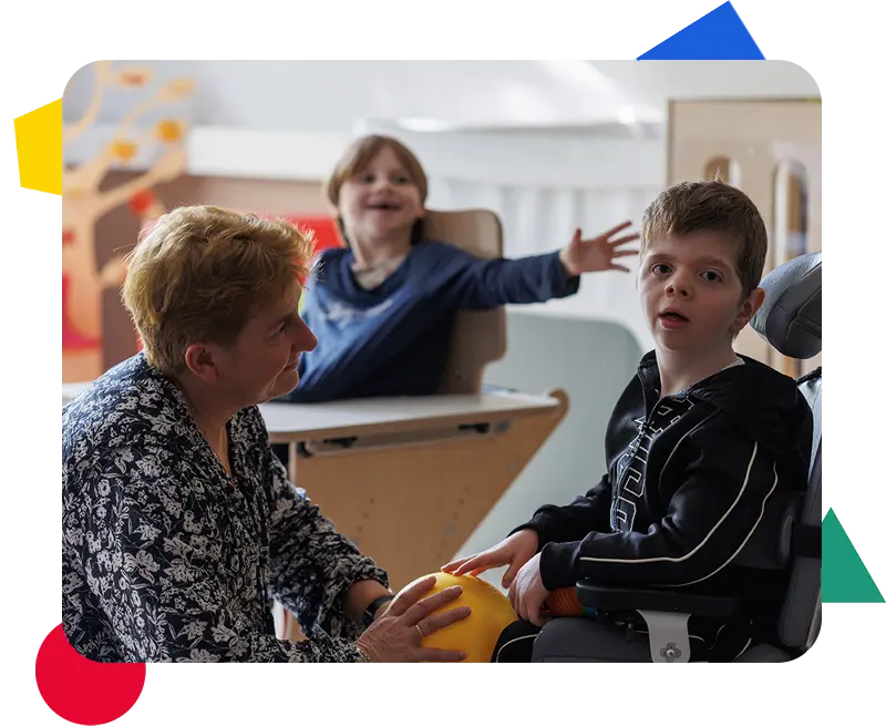 Een vrouw zit tegenover een jongen in een rolstoel in een klaslokaal en houdt een gele bal vast. Op de achtergrond zit een ander kind aan een tafel met uitgestrekte arm.